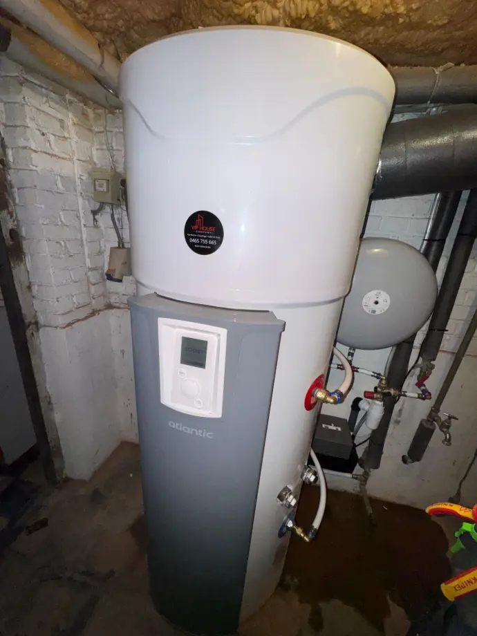 Photo d’un boiler thermodynamique moderne installé dans une buanderie lumineuse. On y voit le réservoir vertical en acier blanc avec son afficheur digital, des conduites d’eau bien organisées et un espace propre et épuré. L’image illustre l’efficacité énergétique et la technologie écologique, idéale pour optimiser la production d’eau chaude tout en réduisant la consommation électrique.